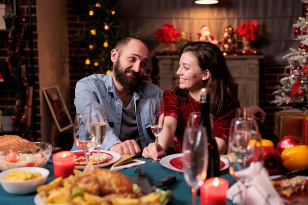 Casal sorridente comemorando o natal em casa, bebendo vinho espumante, esposa e marido segurando copos no jantar festivo. Celebração de Natal com a família, pessoas reunidas à mesa