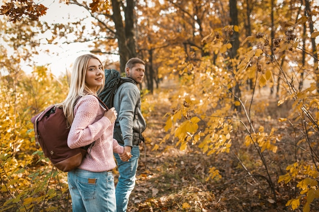 Casal sorridente de turistas andando por um caminho de floresta de mãos dadas e olhando para a câmera