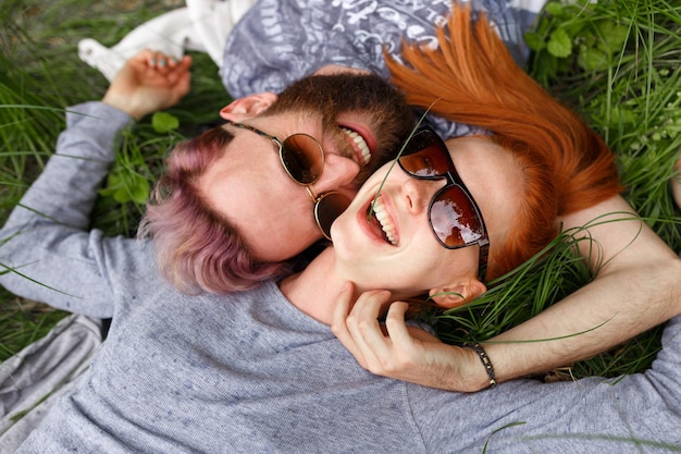 Casal sorridente, lazer em uma grama verde, no parque, sentado com a cabeça virada, de óculos escuros, olhando para a câmera.