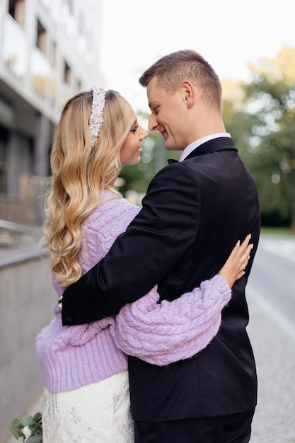 Casal sorridente perfeito de jovem noiva com cabelo louro ondulado em vestido de noiva lilás cardigã em pé hu