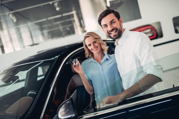 Casal sorridente segurando as chaves em pé no carro novo no salão da concessionária