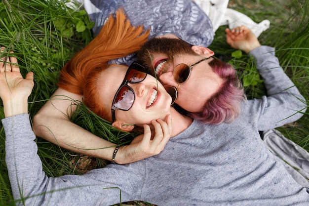 Casal sorridente, sentado em uma grama verde, no parque, sentado com a cabeça voltada, de óculos escuros, olhando para a câmera.