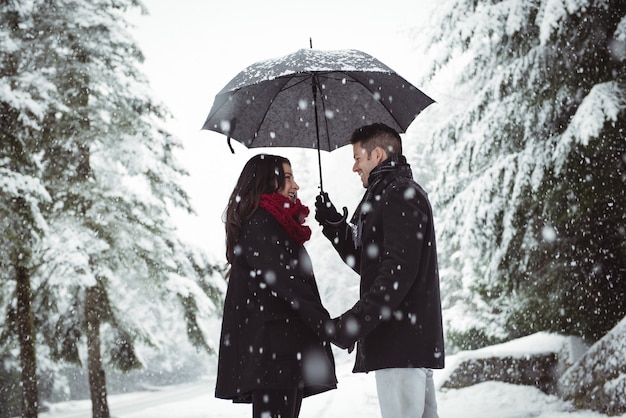 Casal sorridente sob o guarda-chuva em pé na floresta durante a queda de neve