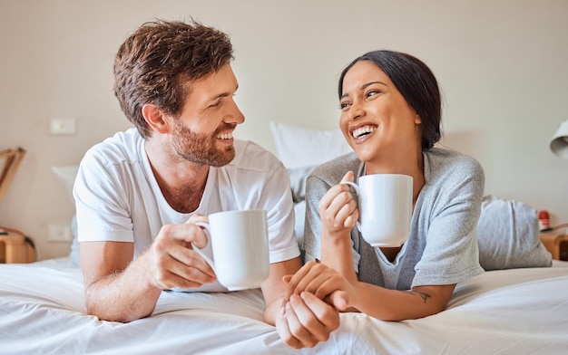 Casal sorriso interracial de café e relaxe na cama em casa sorria para casamento por amor e comunicação engraçada no quarto de casa Homem e mulher felizes com bebida de chá e conversa em quadrinhos pela manhã