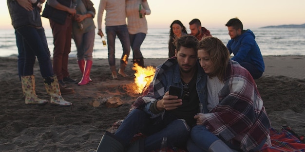 Casal usando celular durante festa na praia com amigos bebendo cerveja e se divertindo