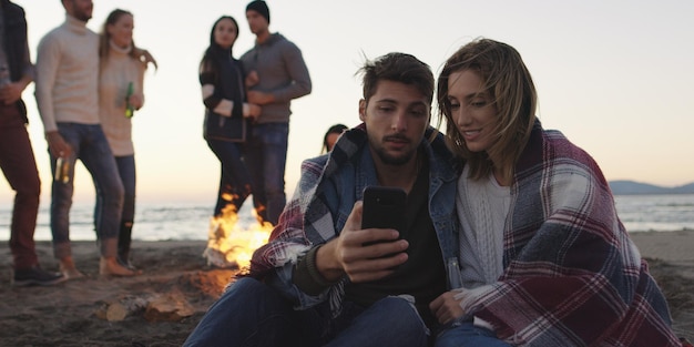 Casal usando celular durante festa na praia com amigos bebendo cerveja e se divertindo