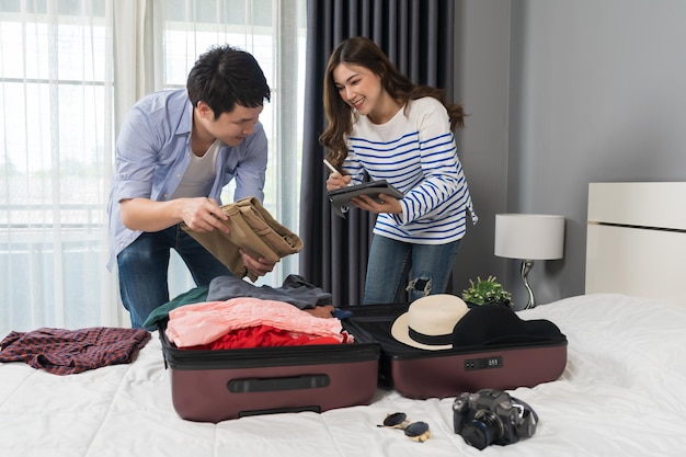 Casal verifica uma lista de coisas com um tablet preparar e embalar roupas na mala na cama planejando férias de viagem