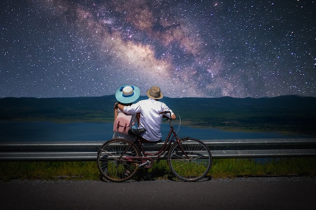 Casal viajante sentado perto de uma bicicleta e olhando para a via láctea e estrelas no céu