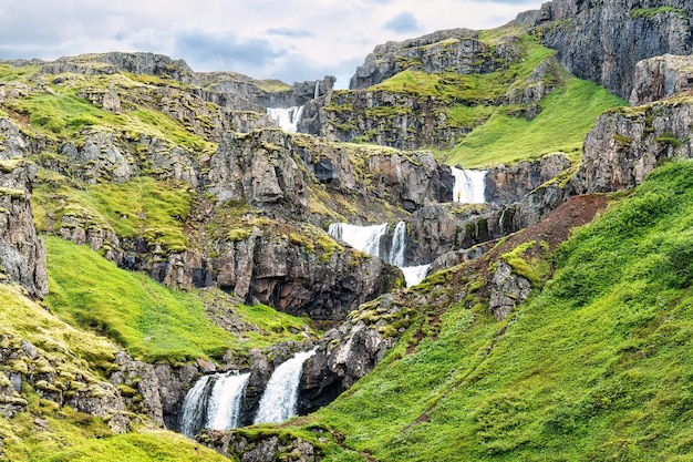 Cascada de varios niveles Klifbrekkufossar que fluye desde un acantilado entre musgo verde en el fiordo Mjoifjordur en verano en Islandia