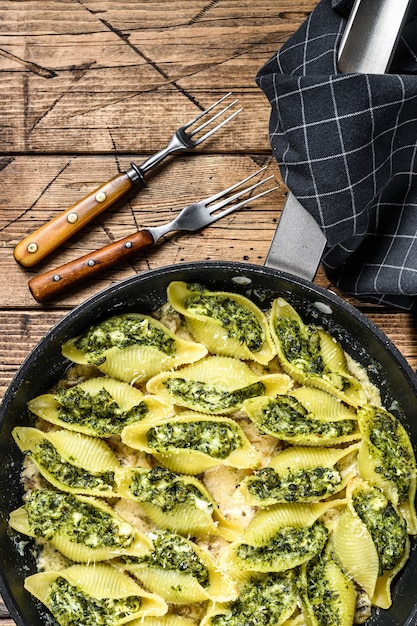 Cascas de massa Conchiglioni recheadas com espinafre e queijo assado com molho em uma panela na mesa de madeira. Vista do topo.