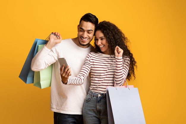 Foto cashback alegre pareja árabe sosteniendo bolsas de compras y celebrando el éxito con el teléfono inteligente