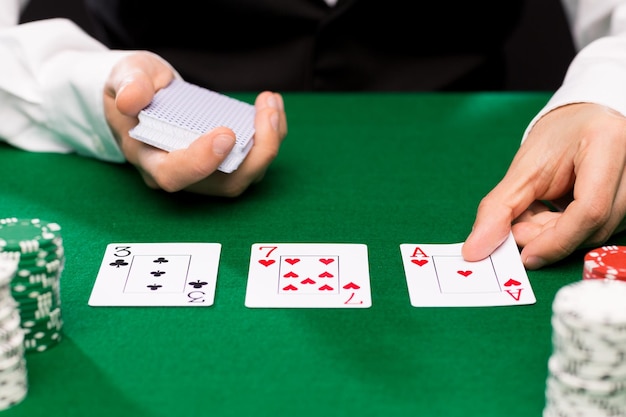 cassino, jogos de azar, pôquer, pessoas e conceito de entretenimento - close-up do negociante de holdem com cartas de baralho e fichas na mesa verde