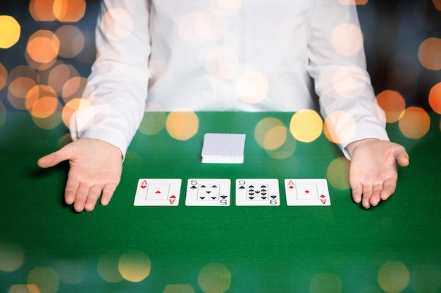 cassino, jogos de azar, pôquer, pessoas e conceito de entretenimento - close-up do negociante de holdem com cartas de baralho na mesa verde sobre fundo de luzes de férias