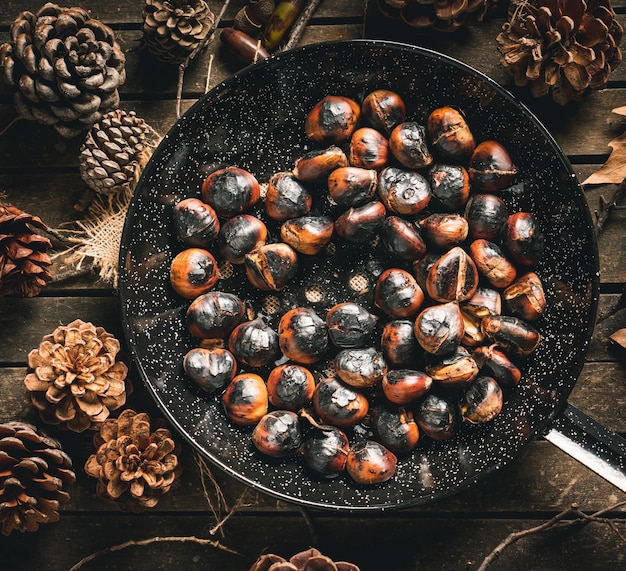 Castanhas assadas em uma frigideira de castanhas com furos em uma mesa com folhas de outono e pinha