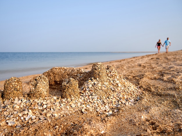 Castillo de arena con conchas en la playa contra el fondo de dos personas cogidas de la mano