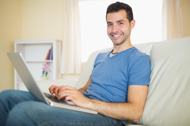 Casual contenido hombre sentado en el sofá con la computadora portátil mirando la cámara