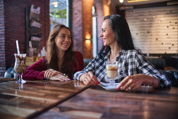 Casual feliz sonriente moderna madre e hija tomando café juntos y disfrutando de la conversación entre ellos en una cafetería