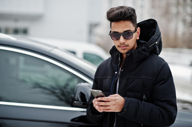 Casual hombre indio joven con chaqueta negra y gafas de sol posó contra suv coche con teléfono en las manos.