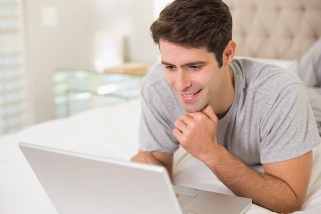 Casual hombre sonriente usando la computadora portátil en la cama