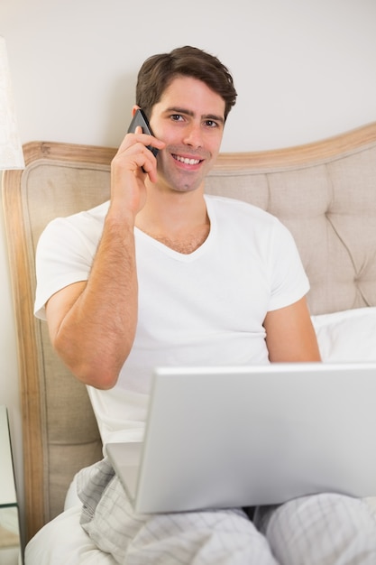 Casual homem sorridente usando celular e laptop na cama