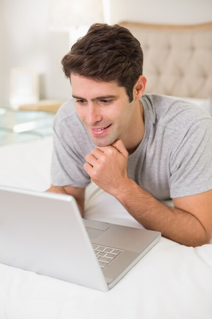 Casual joven sonriente usando la computadora portátil en la cama