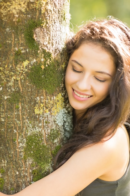 Foto casual morena sonriente abrazando un árbol con los ojos cerrados