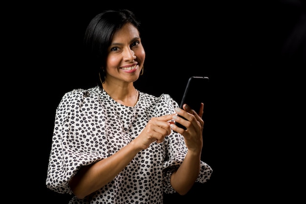 Casual mujer latinoamericana de mediana edad mirando y mostrando un teléfono inteligente sin marca sobre fondo negro