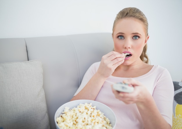 Casual rubia asombrada con control remoto y comer palomitas de maíz