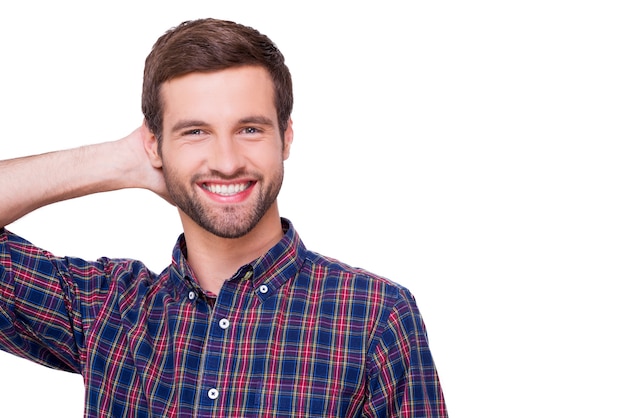 Casualmente guapo. Retrato de joven guapo en camisa casual sosteniendo la mano detrás de la cabeza y sonriendo mientras está de pie aislado en blanco