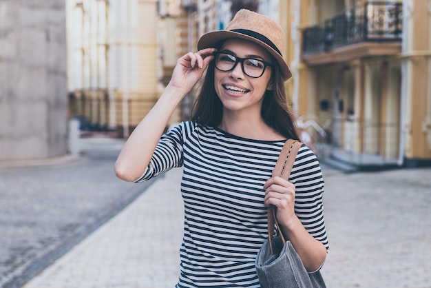 Casualmente hermosa. Hermosa mujer joven con bolsa sobre el hombro mirando a la cámara y sonriendo mientras está de pie al aire libre