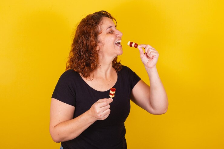 Foto caucasiana brasileira latino-americana cabelos cacheados fundo amarelo sorrindo segurando espetos de queijo e goiaba surpreso rosto romeu e julieta doce delicioso
