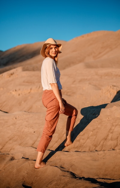 Caucasiana linda jovem sozinha está em uma gostosa. Pôr do sol no deserto. Menina andando na areia dourada.