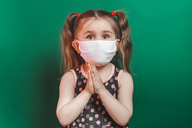 Caucasiana menina doente na máscara médica durante a epidemia de coronavírus reza sobre fundo verde closeup.