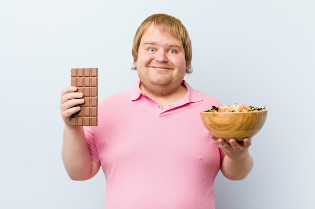 Caucasiano louco gordo loiro escolhendo entre tablete de chocolate ou saladeira