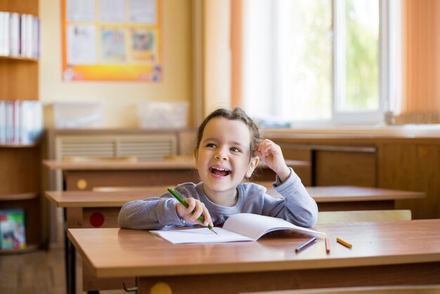 Caucásica niña sonriente sentada en el escritorio en la sala de clase y comienza a dibujar cuidadosamente en un cuaderno puro.