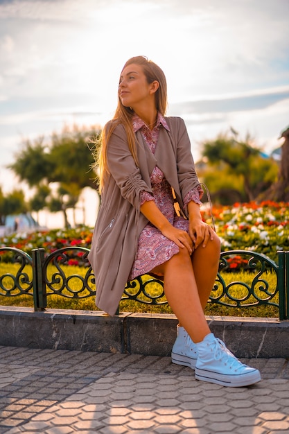 Caucásica y rubia con impermeable marrón y vestido de flores en la ciudad. Chica joven atractiva con el sol en el fondo junto a flores en un hermoso parque