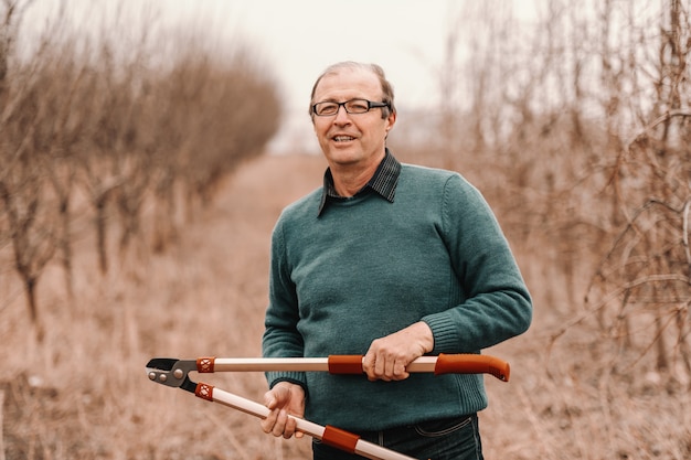 Caucásico agrónomo senior con anteojos con tijeras de poda y de pie en el huerto.