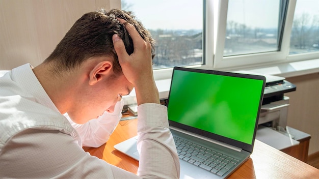 Un caucásico frustrado sostiene su cabeza mientras se sienta en una mesa frente a una computadora con una s verde