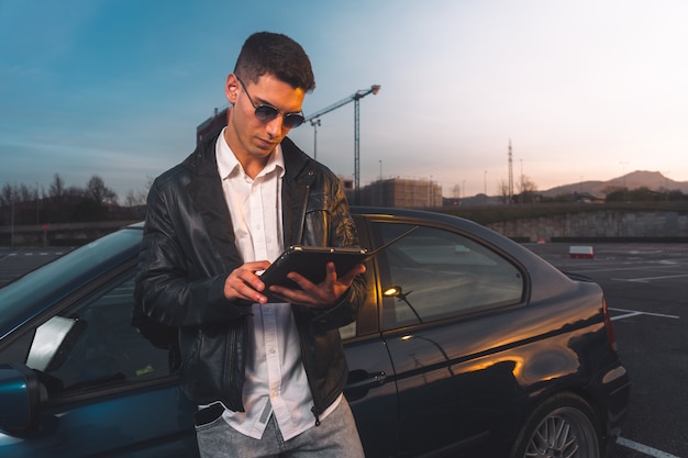 Caucásico joven usando una tableta digital con un coche deportivo detrás en un estacionamiento.