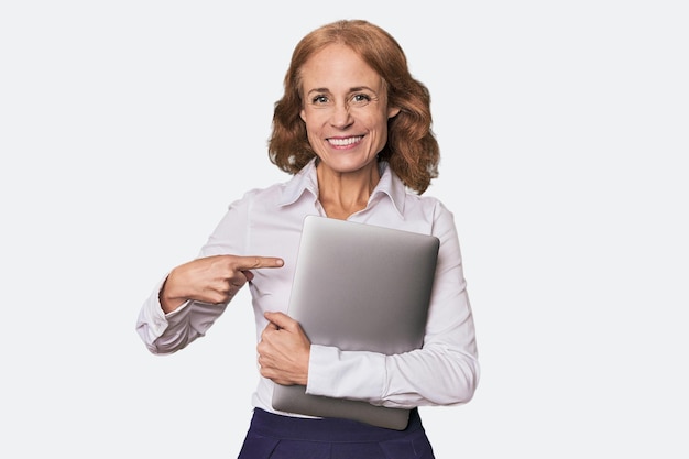 Caucaso de mediana edad con computadora portátil en el estudio sonriendo y señalando a un lado mostrando algo en espacio en blanco