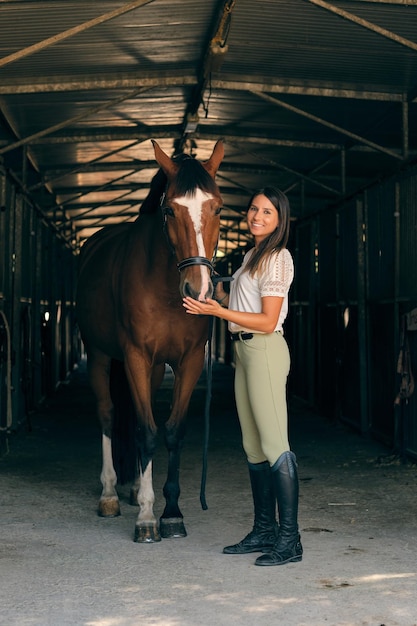 Cavaleira encantada de pé com um cavalo obediente castanho no celeiro no campo e olhando para a câmera