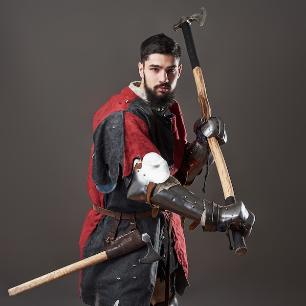 Cavaleiro medieval em fundo cinza. retrato de guerreiro de rosto brutal com roupa vermelha e preta de armadura de cota de malha e machado de batalha.