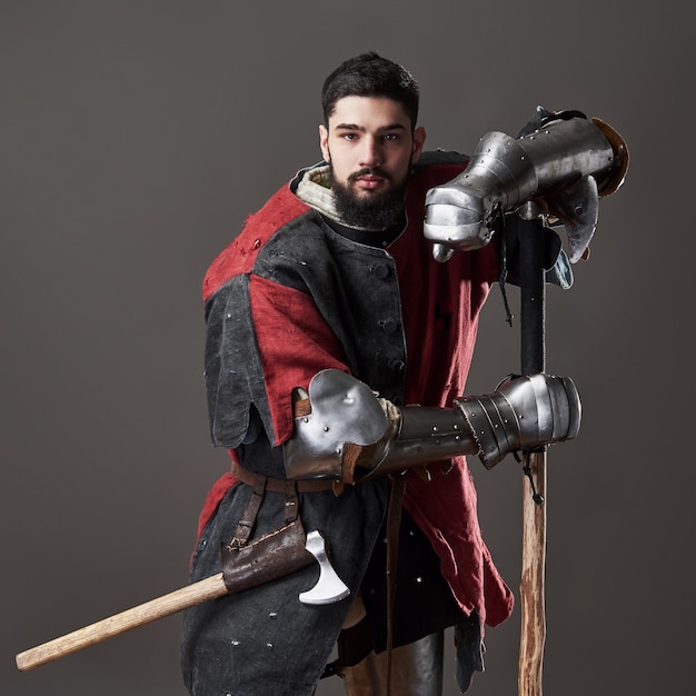 Foto cavaleiro medieval em fundo cinza. retrato de guerreiro de rosto brutal com roupa vermelha e preta de armadura de cota de malha e machado de batalha.