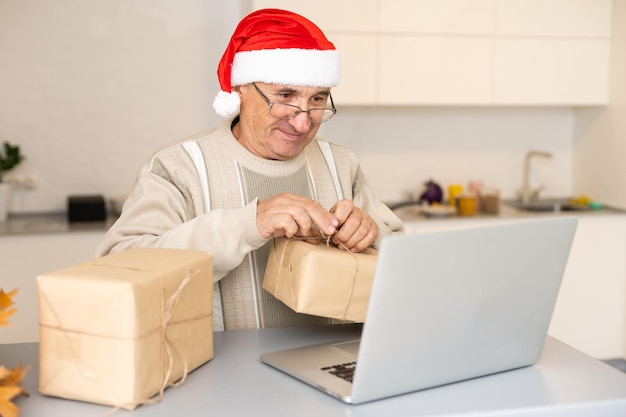 Cavalheiro idoso celebrando o Natal on-line no laptop fazendo videochamada com a família distante sentada na cozinha em casa. Feriado de ano novo.