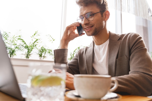 Cavalheiro sorridente de óculos sentado à mesa com um laptop e conversando ao telefone