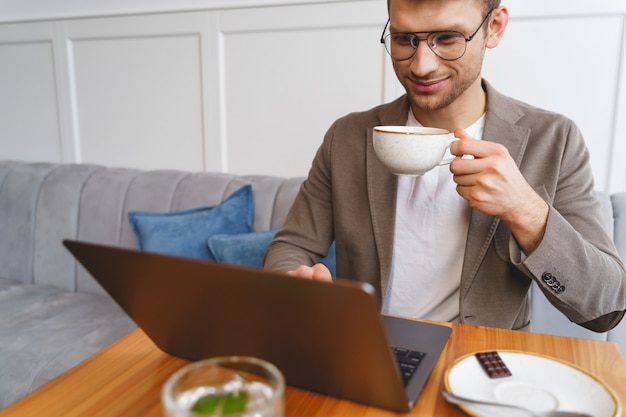 Cavalheiro sorridente segurando um copo de bebida quente enquanto está sentado à mesa com um laptop moderno no café