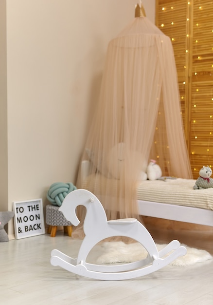 Cavalo de madeira branco no interior de um quarto de criança fofo com brinquedos e móveis modernos, cama com tenda