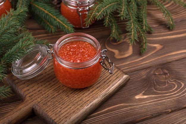 Caviar rojo en un frasco de vidrio sobre un fondo de madera marrón con un árbol de Navidad.