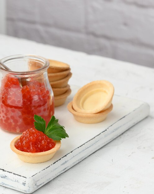 Foto caviar rojo en un frasco de vidrio y tortillas redondas en una mesa blanca de cerca