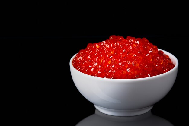 Caviar rojo en una taza blanca sobre un fondo negro con un reflejo. Lugar para publicidad, logotipo, etiqueta, maqueta, maqueta.